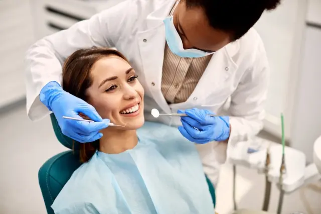 Mulher sorrindo na cadeira odontológica enquanto profissional de saúde usa espelho e explorador com luvas azuis