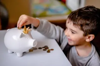 Criança sorrindo colocando uma moeda em um cofrinho branco em formato de porquinho com uma pequena coroa dourada, com moedas espalhadas sobre a mesa
