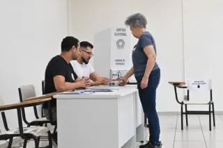 Eleitora em pé entregando documento a mesários sentados junto a uma mesa de votação