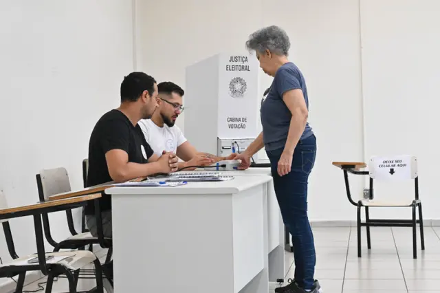 Eleitora em pé entregando documento a mesários sentados junto a uma mesa de votação