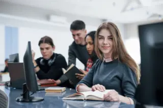 Jovem mulher sorrindo com livro aberto à frente de monitores, colegas ao fundo em ambiente com computadores