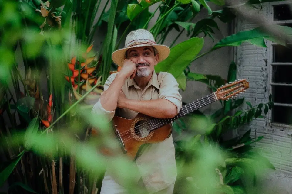 homem de chapéu segurando uma viola entre plantas verdes, com folhas desfocadas em primeiro plano