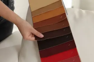 mão segurando catálogo de amostras com tiras de tecido e couro em tons do bege ao vermelho sobre um sofá branco