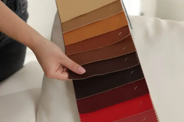 mão segurando catálogo de amostras com tiras de tecido e couro em tons do bege ao vermelho sobre um sofá branco