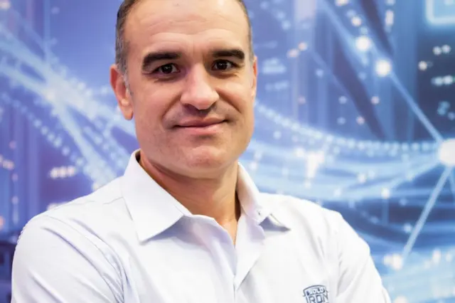 Homem de meia-idade de braços cruzados vestindo camisa branca com logotipo Solo Iron, em frente a um fundo azul com elementos gráficos tecnológicos