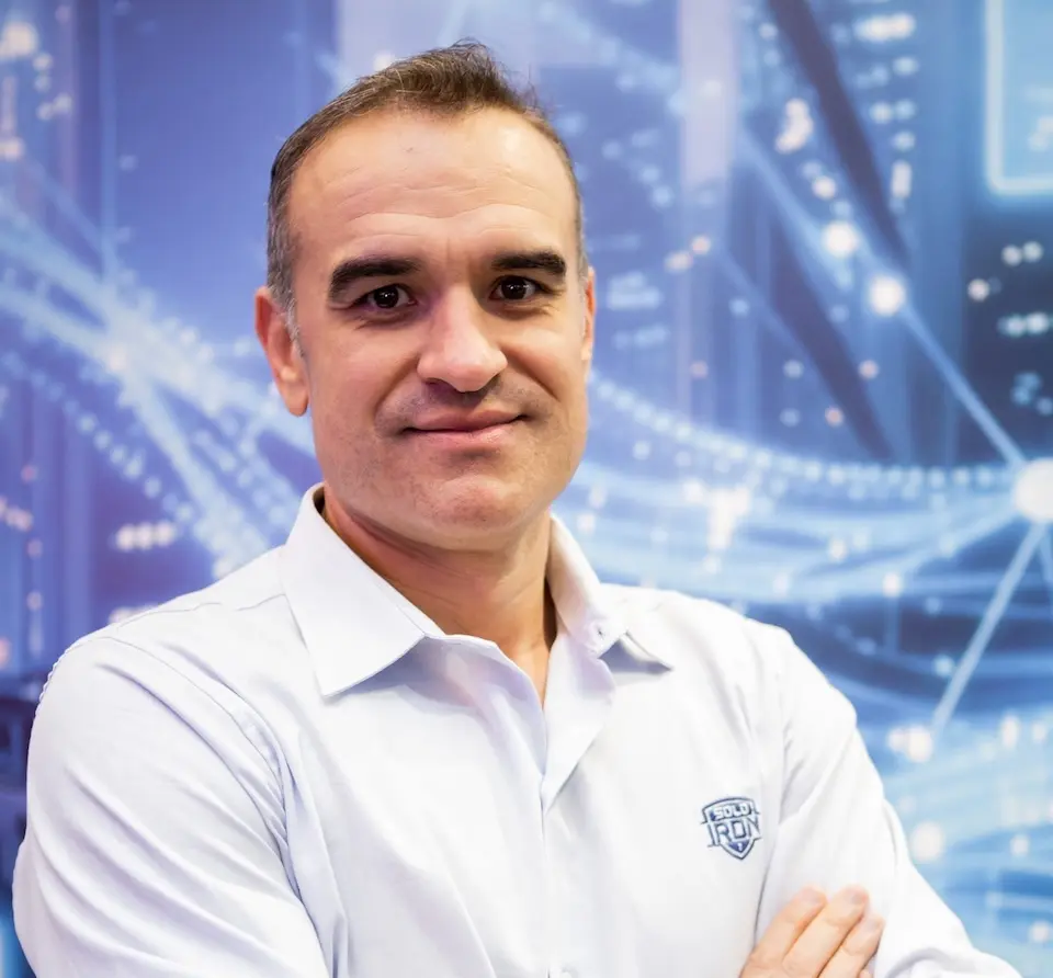 Homem de meia-idade de braços cruzados vestindo camisa branca com logotipo Solo Iron, em frente a um fundo azul com elementos gráficos tecnológicos