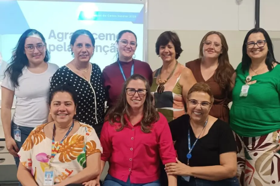 Nove mulheres adultas sorrindo em duas fileiras em sala com tela de projeção ao fundo