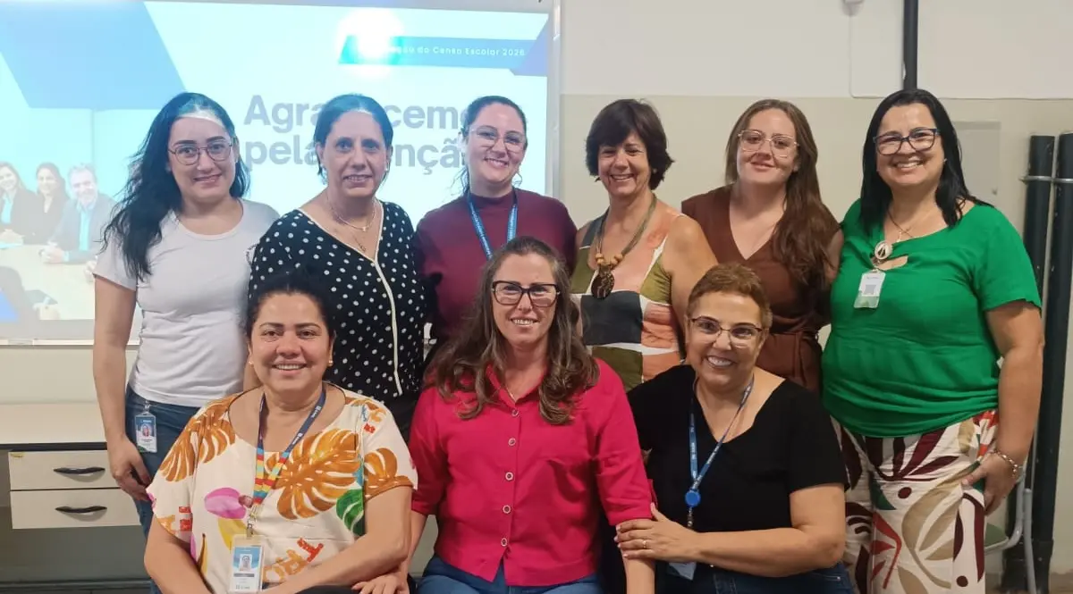 Nove mulheres adultas sorrindo em duas fileiras em sala com tela de projeção ao fundo