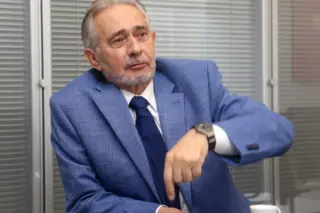 homem idoso de pele clara, cabelo e barba grisalhos, vestindo paletó azul e gravata escura, sentado à mesa apontando com a mão direita; xícara de café sobre a mesa; fundo com persianas