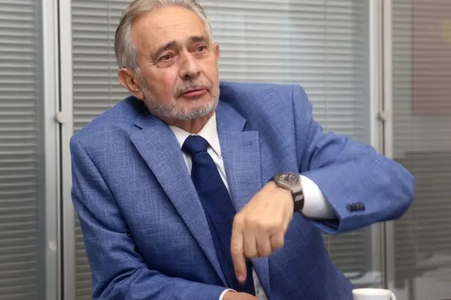homem idoso de pele clara, cabelo e barba grisalhos, vestindo paletó azul e gravata escura, sentado à mesa apontando com a mão direita; xícara de café sobre a mesa; fundo com persianas