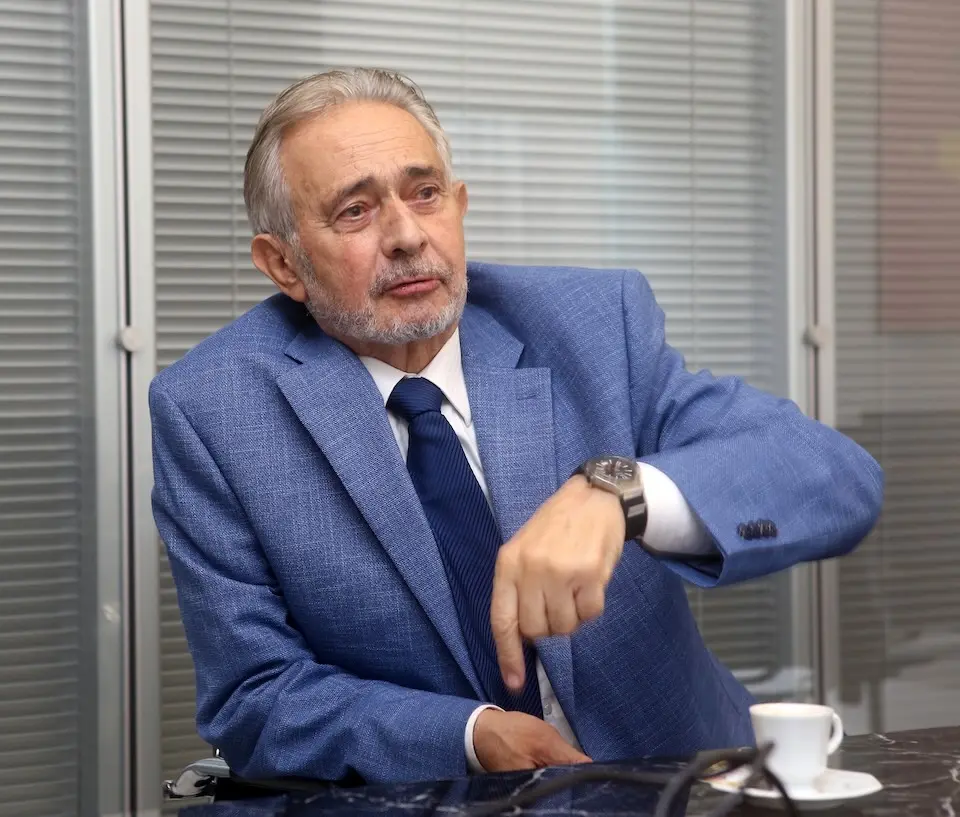 homem idoso de pele clara, cabelo e barba grisalhos, vestindo paletó azul e gravata escura, sentado à mesa apontando com a mão direita; xícara de café sobre a mesa; fundo com persianas