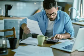 Homem adulto de óculos e barba sentado à mesa usando uma calculadora, com papéis, laptop e caneca ao redor