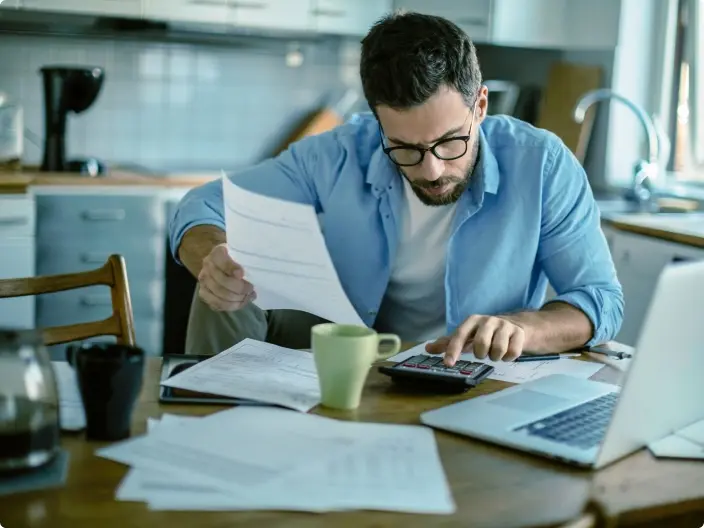 Homem adulto de óculos e barba sentado à mesa usando uma calculadora, com papéis, laptop e caneca ao redor