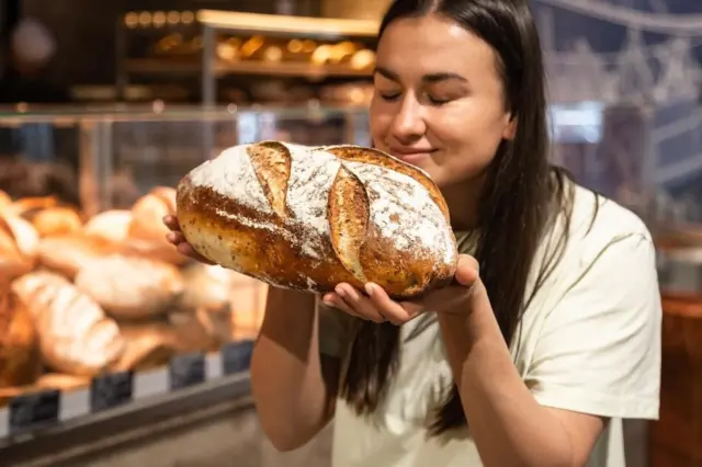 Mulher segurando e cheirando um pão grande em padaria com prateleira de pães ao fundo