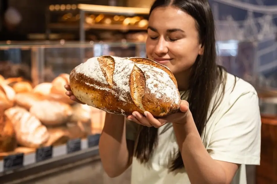 Mulher segurando e cheirando um pão grande em padaria com prateleira de pães ao fundo
