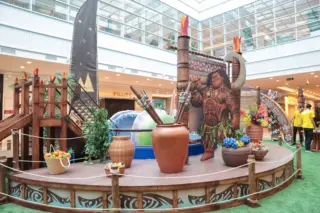 cenário temático infantil em área interna de shopping com grande figura do personagem Maui, estrutura de madeira, vasos, piscina de bolinhas e piso em grama sintética