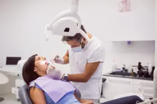 dentista com máscara e luvas examinando a boca de uma paciente mulher reclinada na cadeira odontológica