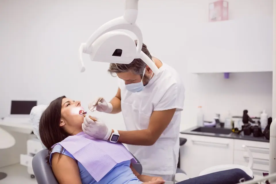 dentista com máscara e luvas examinando a boca de uma paciente mulher reclinada na cadeira odontológica