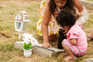 Duas meninas em um cemitério: uma mais velha inclinada tocando uma lápide e outra menor agachada ao lado, com uma lanterna e flores sobre a sepultura