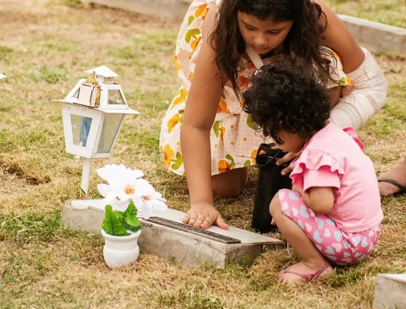 Duas meninas em um cemitério: uma mais velha inclinada tocando uma lápide e outra menor agachada ao lado, com uma lanterna e flores sobre a sepultura