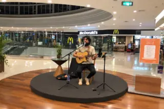 Cantor sentado em palco circular tocando violão na praça de alimentação do shopping, com lanchonete McDonald's ao fundo