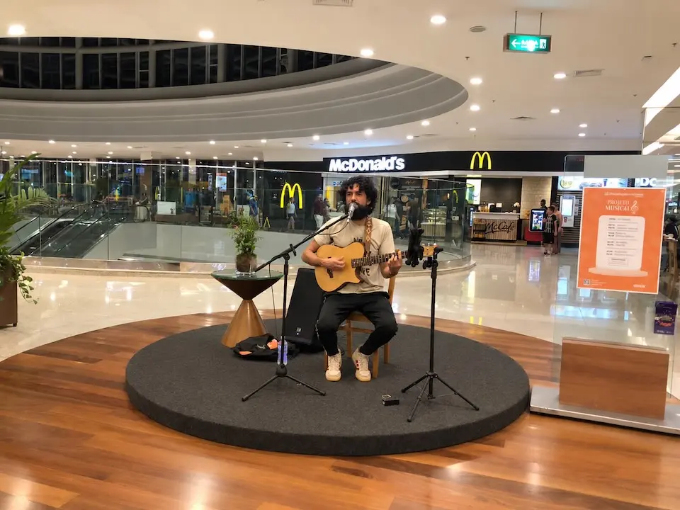 Cantor sentado em palco circular tocando violão na praça de alimentação do shopping, com lanchonete McDonald's ao fundo