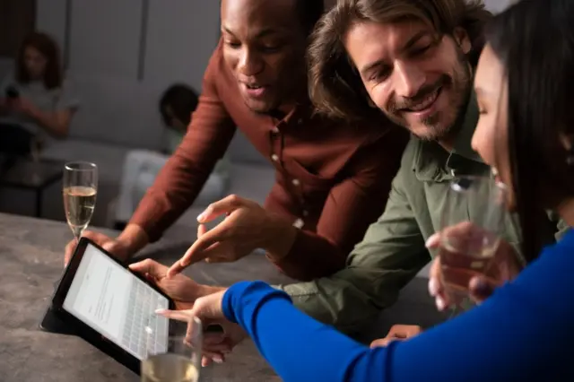 Três pessoas reunidas apontando e vendo conteúdo em um tablet sobre uma bancada, com taças de bebida ao redor