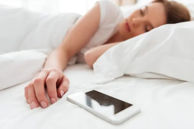 mulher dormindo deitada na cama com a mão estendida próxima a um smartphone sobre o lençol branco