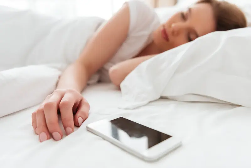mulher dormindo deitada na cama com a mão estendida próxima a um smartphone sobre o lençol branco