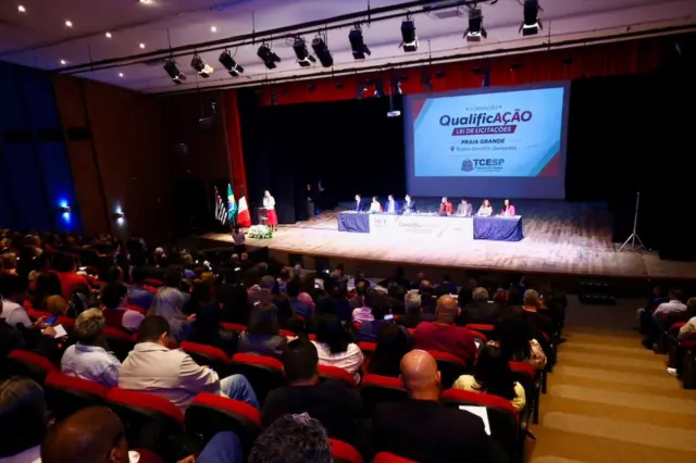 Auditório com plateia e palco onde há mesa de palestrantes e tela de projeção com o logotipo do TCESP e a palavra 'QualificAÇÃO'.