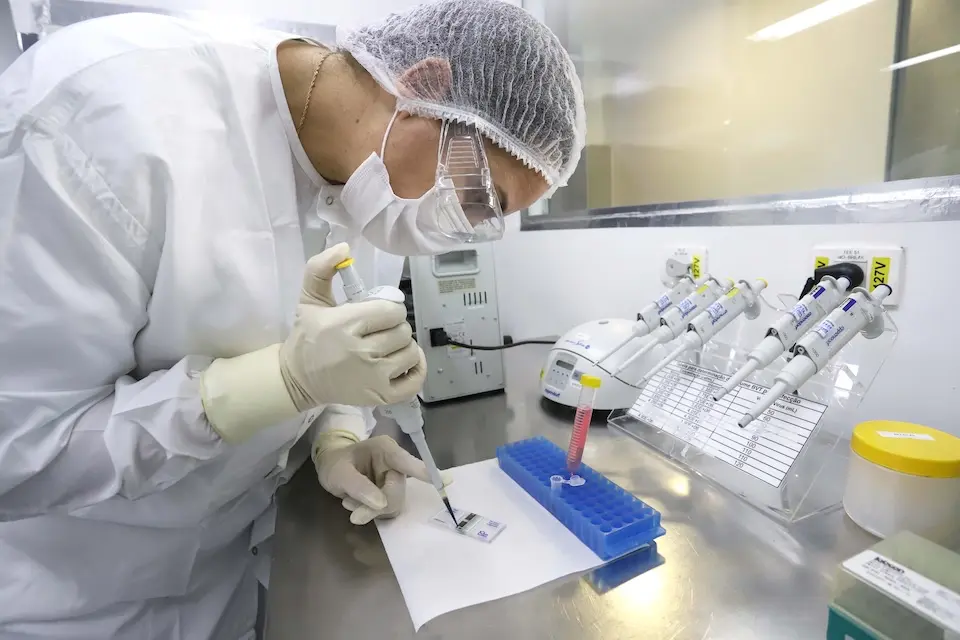 Técnico de laboratório com touca, máscara, óculos e jaleco using micropipeta sobre lâmina em bancada com rack azul, microtubo com tampa amarela e micropipetas ao fundo
