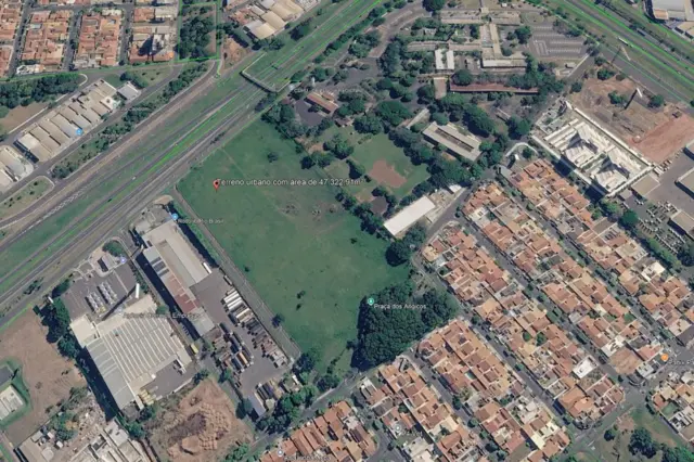 Terreno gramado amplo visto em imagem aérea, ladeado por via expressa, galpões industriais e bairro residencial