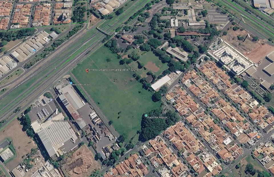 Terreno gramado amplo visto em imagem aérea, ladeado por via expressa, galpões industriais e bairro residencial