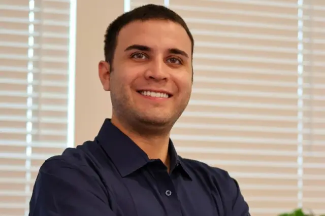 Homem sorridente com os braços cruzados, usando camisa azul escura, persianas brancas atrás e planta ao fundo