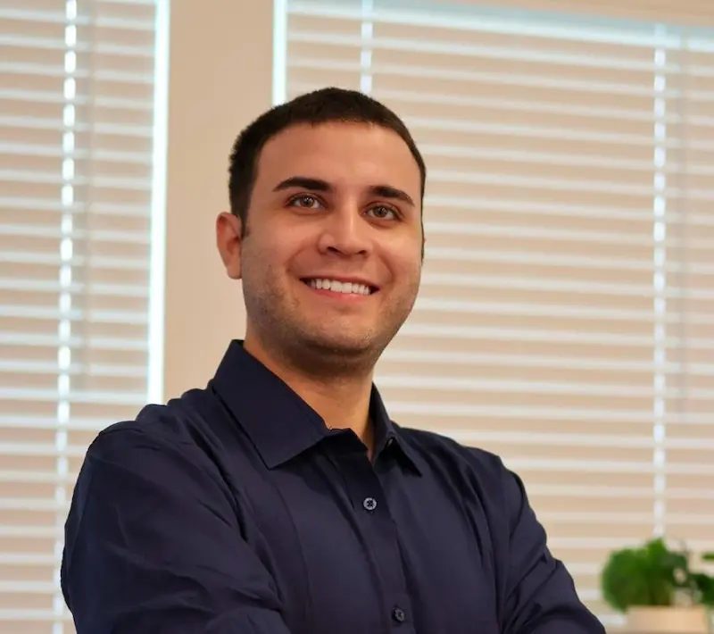 Homem sorridente com os braços cruzados, usando camisa azul escura, persianas brancas atrás e planta ao fundo