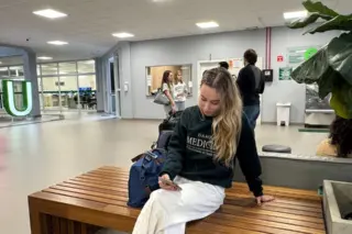 mulher jovem sentada em banco de madeira em saguão universitário olhando para o celular, com mochila ao lado e pessoas ao fundo