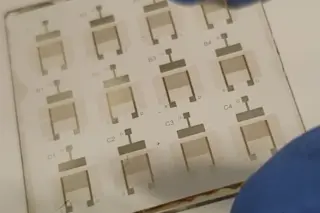 Placa com padrões impressos de transistores em forma de U e marcações alfanuméricas, com luva azul visível no canto inferior direito