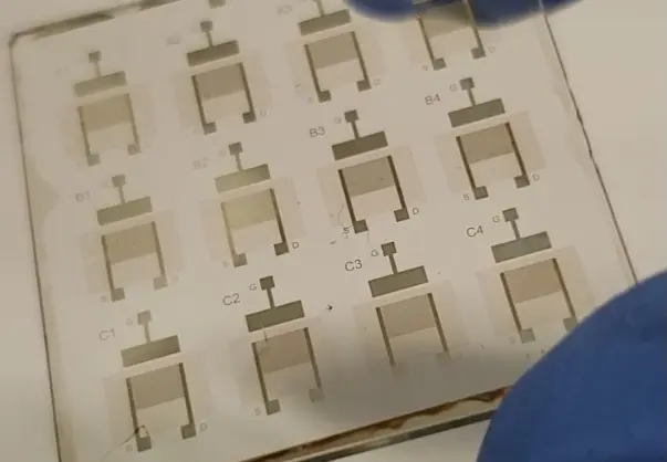 Placa com padrões impressos de transistores em forma de U e marcações alfanuméricas, com luva azul visível no canto inferior direito