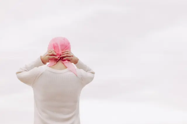 Pessoa vista de costas ajustando lenço rosa na cabeça, vestindo suéter claro sobre fundo claro