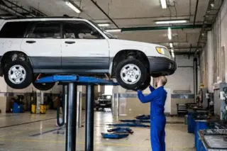 SUV branco elevado em plataforma hidráulica em oficina, com mecânico de macacão azul inspecionando a parte dianteira