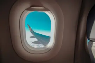 Asa de avião vista por janela de cabine, céu azul e nuvens abaixo