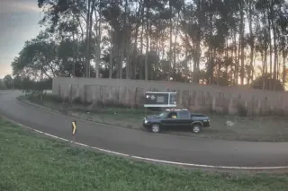 caminhonete estacionada no acostamento de uma via curva, com painel publicitário e muro ao fundo e árvores altas