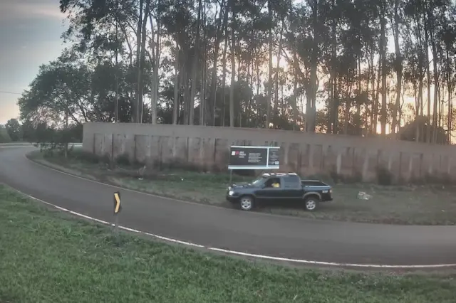 caminhonete estacionada no acostamento de uma via curva, com painel publicitário e muro ao fundo e árvores altas
