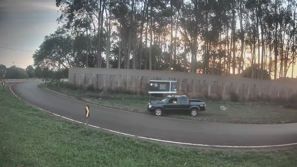 caminhonete estacionada no acostamento de uma via curva, com painel publicitário e muro ao fundo e árvores altas