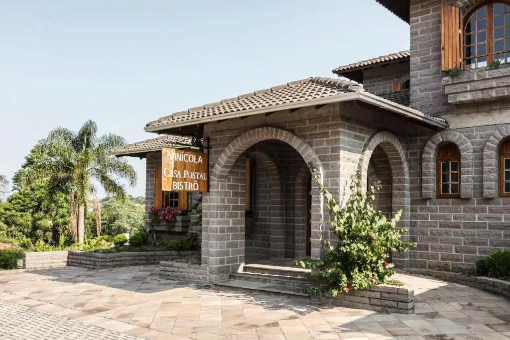 fachada de pedra com arco de entrada e placa de madeira com texto 'VINICOLA Casa Postal BISTRÔ', plantas próximas