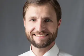 homem adulto sorrindo, barba e cabelo castanho, vestindo camisa social branca e gravata listrada, fundo cinza