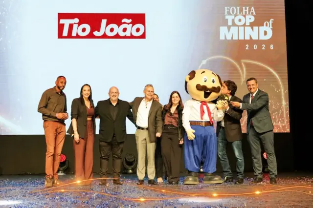 Pessoas em pé no palco ao lado do mascote do Tio João, com o logotipo Tio João e o texto Folha Top of Mind projetados ao fundo