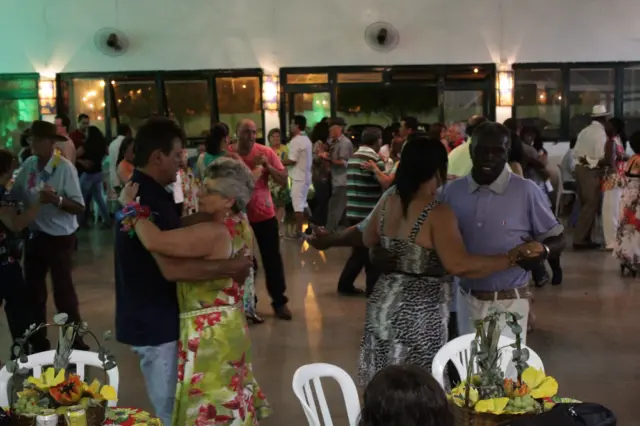 Casais da terceira idade dançando em salão decorado com mesas e arranjos tropicais
