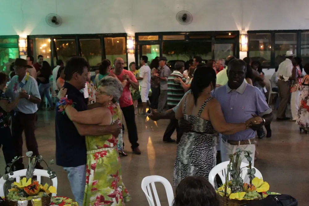 Casais da terceira idade dançando em salão decorado com mesas e arranjos tropicais