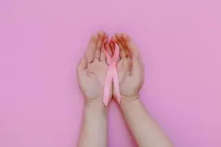 duas mãos abertas em cuia segurando um laço rosa sobre fundo rosa uniforme